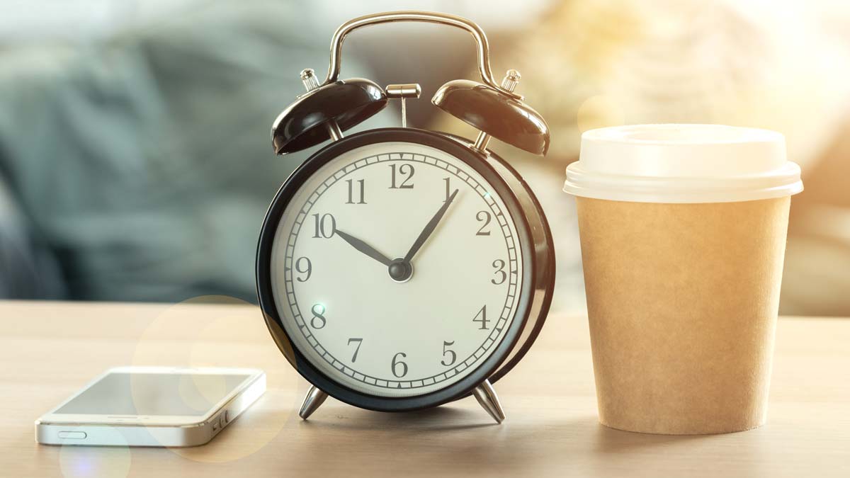 An old fashioned alarm clock reads 5 past 10, there is a cup of coffee and a phone alongside it.